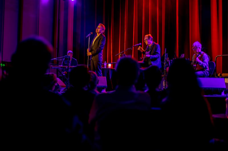 Musicians perform on stage bathed in purple and red lighting, with audience silhouettes in foreground.