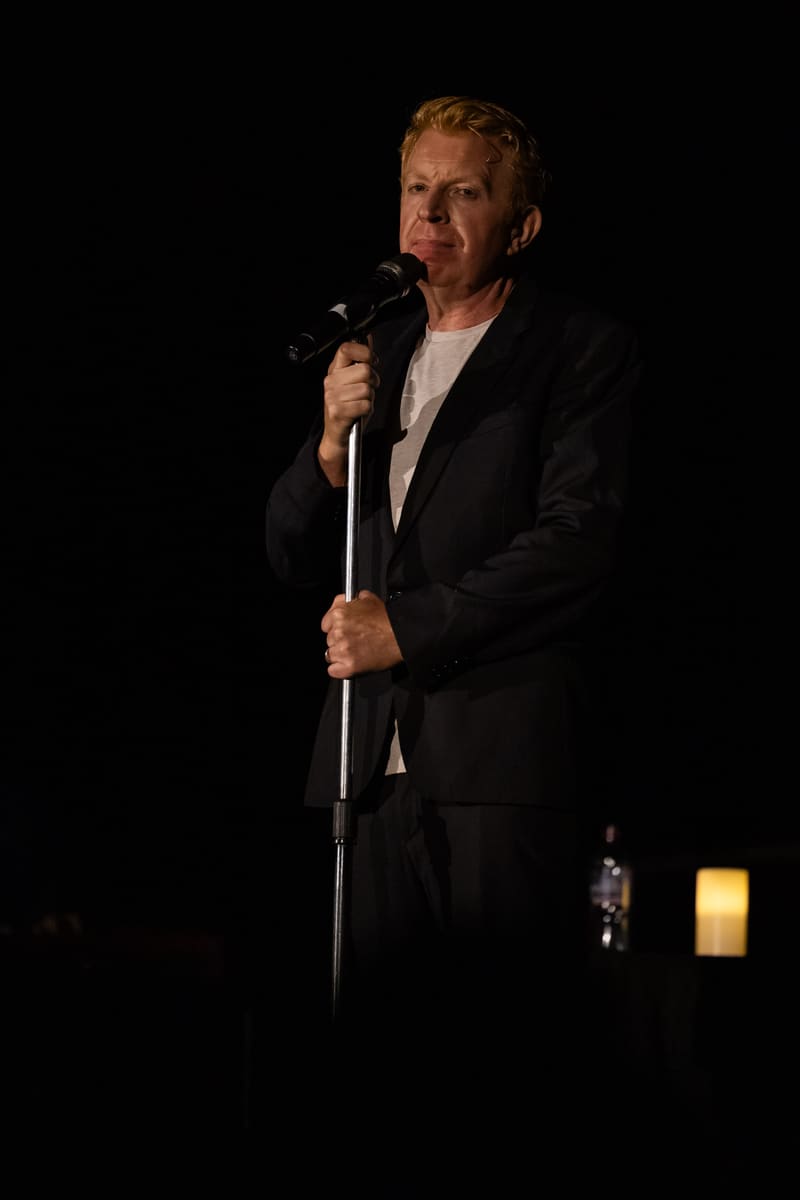 Man in black suit holding microphone stand against dark background, warm stage lighting visible.