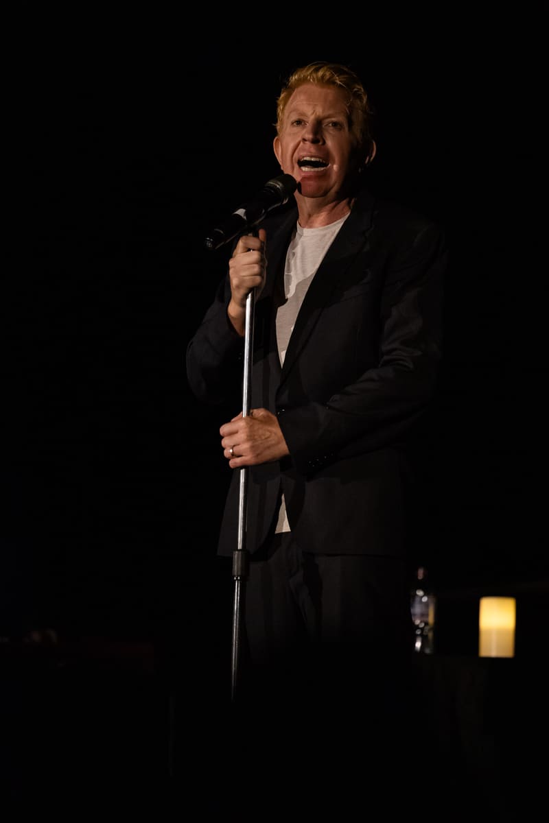 Singer in black suit holding microphone, performing against a dark background with warm stage lighting.