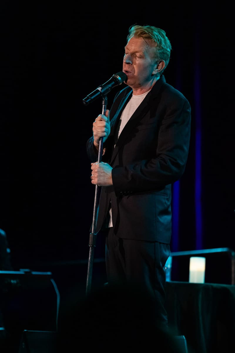 Man in a black suit singing at a microphone on a darkened stage with blue lighting.