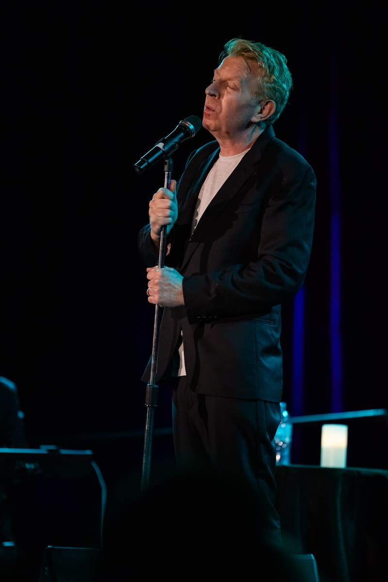 Male singer in black jacket performing at microphone under blue stage lighting.