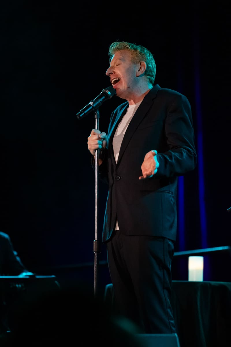 Singer in black suit performing at microphone under blue stage lighting.