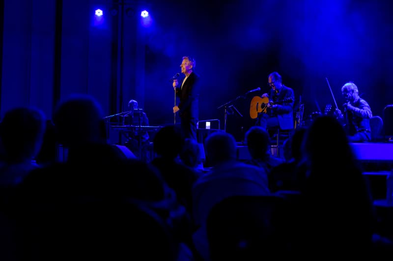 Singer in black suit performs on stage with band members and acoustic guitar, bathed in blue stage lighting.