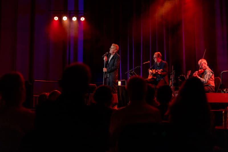 Singer performing centre stage with band members under red and blue stage lighting, audience silhouetted in foreground.