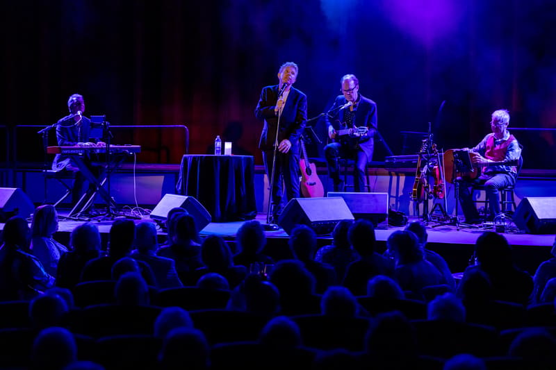 Five musicians perform on a dimly lit stage with blue and purple lighting, silhouettes of audience members visible in the foreground.