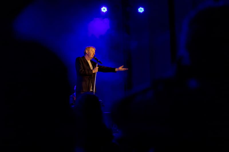 Male performer in dark jacket singing at microphone beneath blue stage lighting.