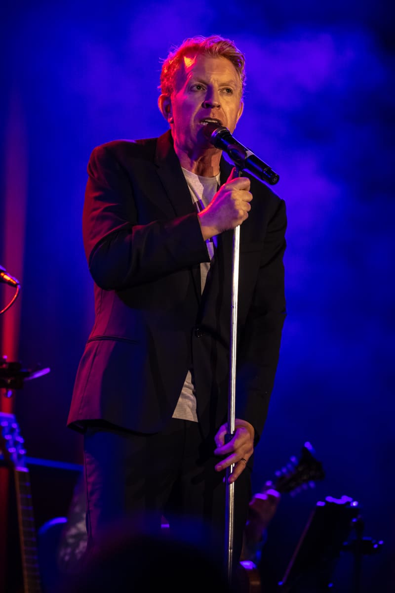 Male vocalist in dark suit performing at microphone under purple and red stage lighting.