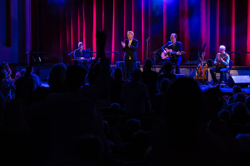 Four musicians perform on stage bathed in blue and magenta lighting, with audience silhouettes in the foreground.