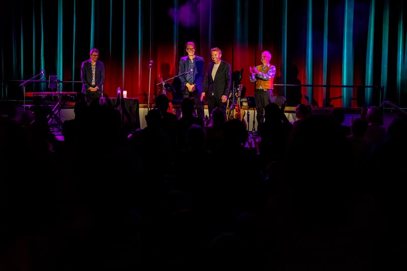 Musicians perform on stage bathed in red and blue lighting with vertical neon tubes behind them.