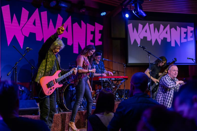 Musician in patterned jacket plays electric guitar on stage beneath Wa Wa Nee signage, with band members and audience visible.