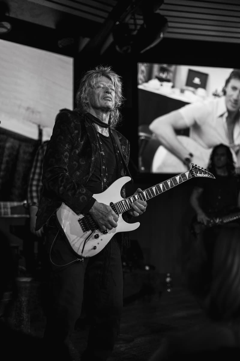 Guitarist performs on stage in black and white, holding an electric guitar under moody stage lighting.