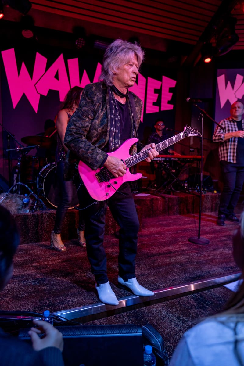 Guitarist performs on stage under pink and purple lighting with "Wa Wa Nee" signage visible behind.