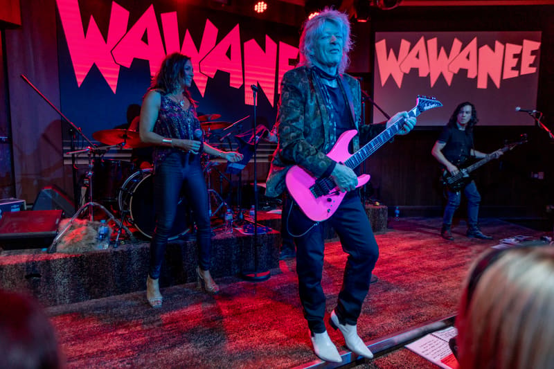 Musician performs with pink electric guitar on red-lit stage beneath Wa Wa Nee signage, flanked by band members.