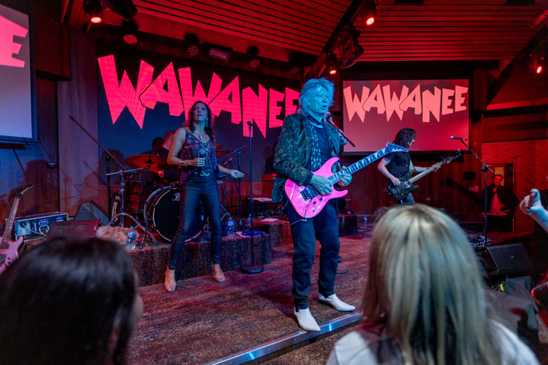 Guitarist performing on stage with neon Wa Wa Nee signage, audience members visible in foreground.