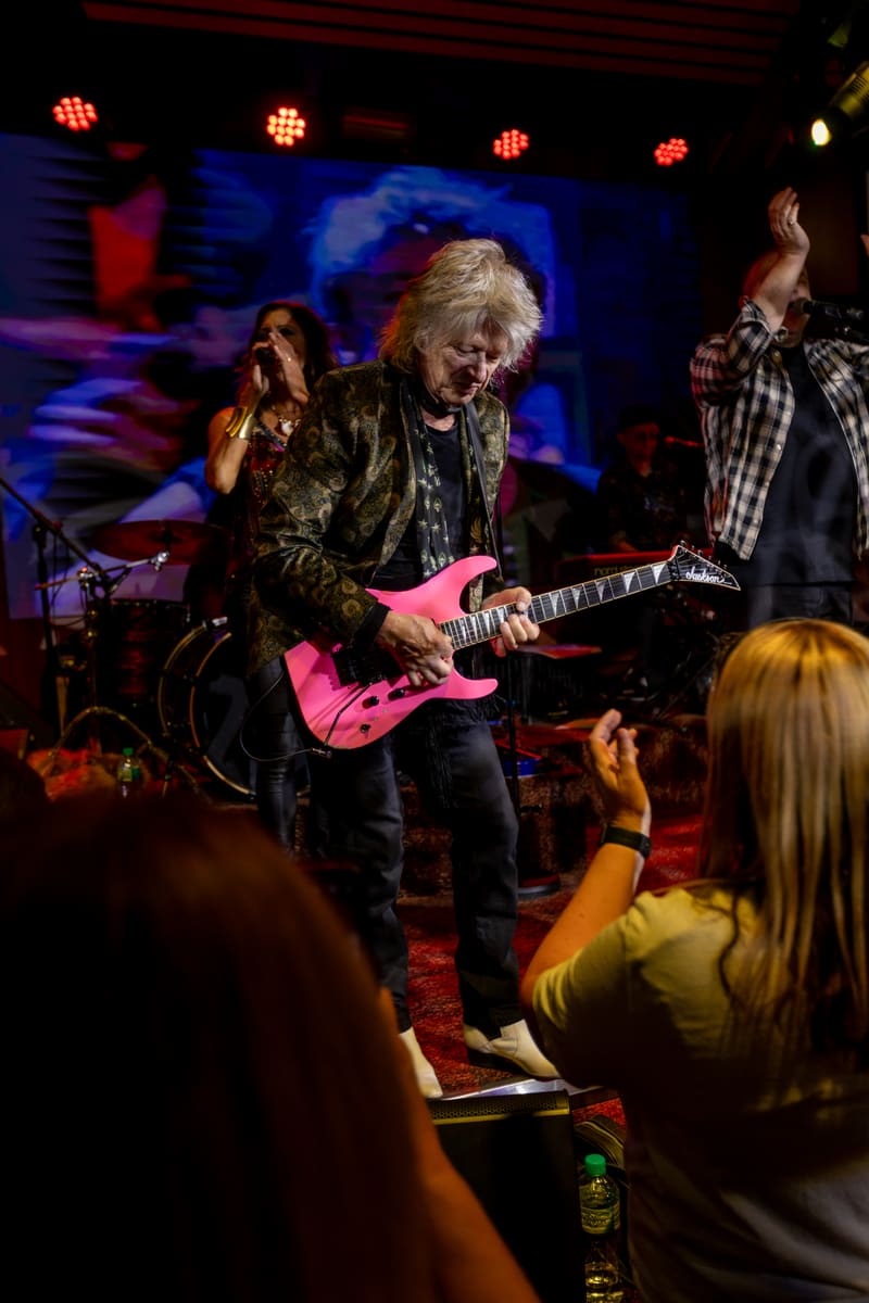 Guitarist performs on a bright pink electric guitar under blue and red stage lights, with audience members in foreground.