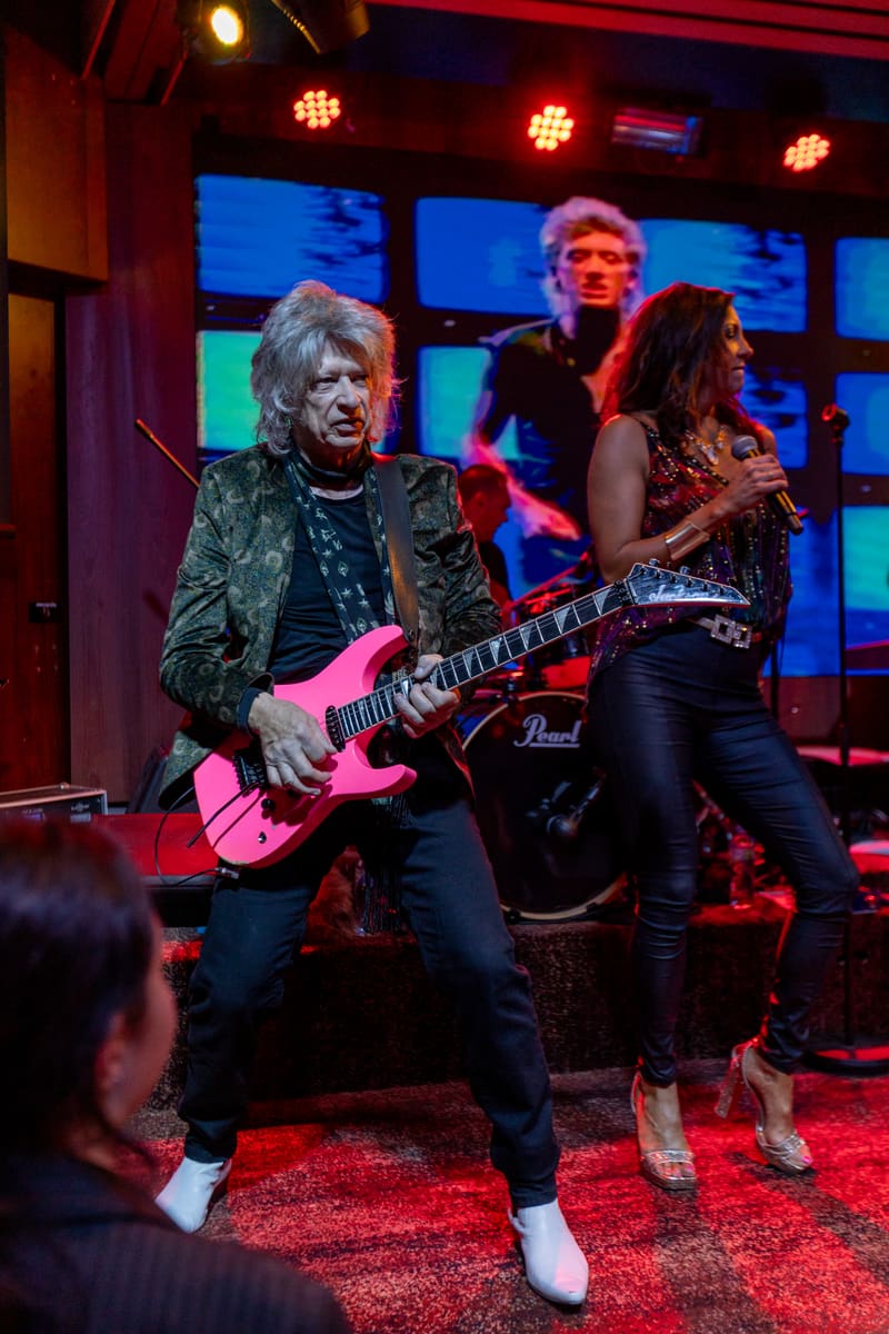 Guitarist in pink-lit stage performance with band members and colourful screen backdrop behind.