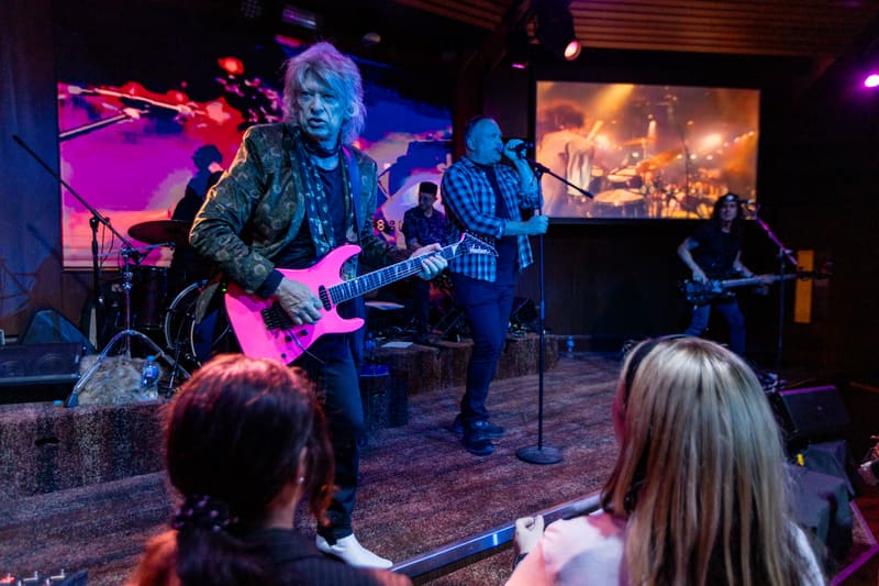 Guitarist in patterned shirt performs centre stage under pink and purple lighting with band members and audience visible.