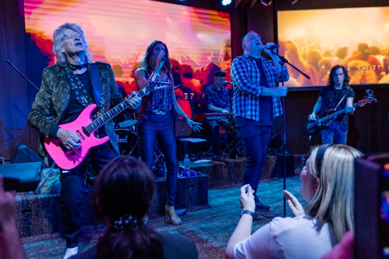 Band members perform on stage under coloured lighting with audience members in the foreground.