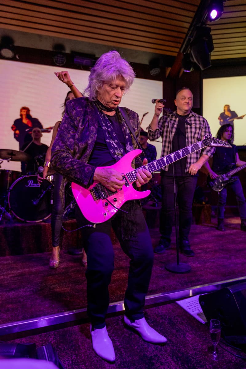 Guitarist in a shimmering purple jacket performs on a bright pink electric guitar under purple stage lighting.