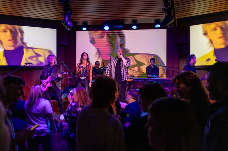 Band performing on stage with audience silhouettes in foreground, purple stage lighting and large screens displaying performers above.