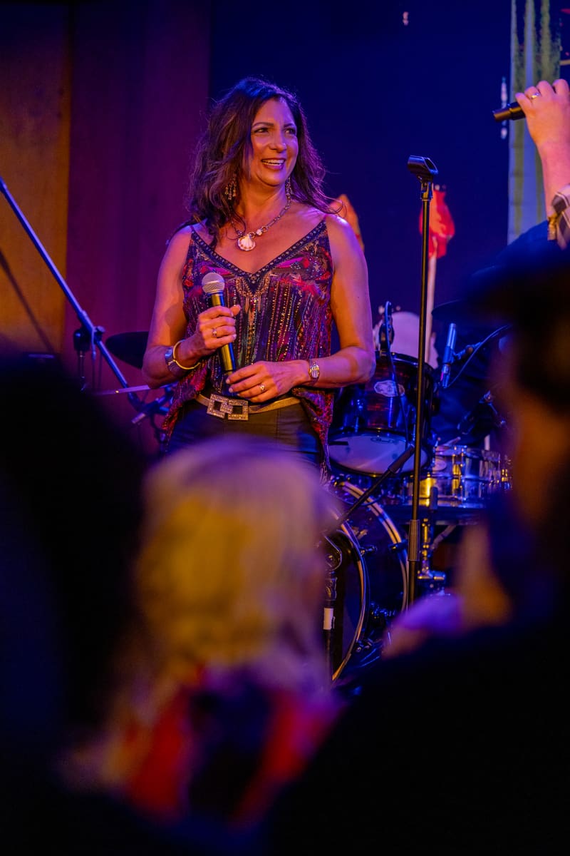 Singer performing on stage in a patterned vest and gold belt, holding a microphone under purple stage lighting.