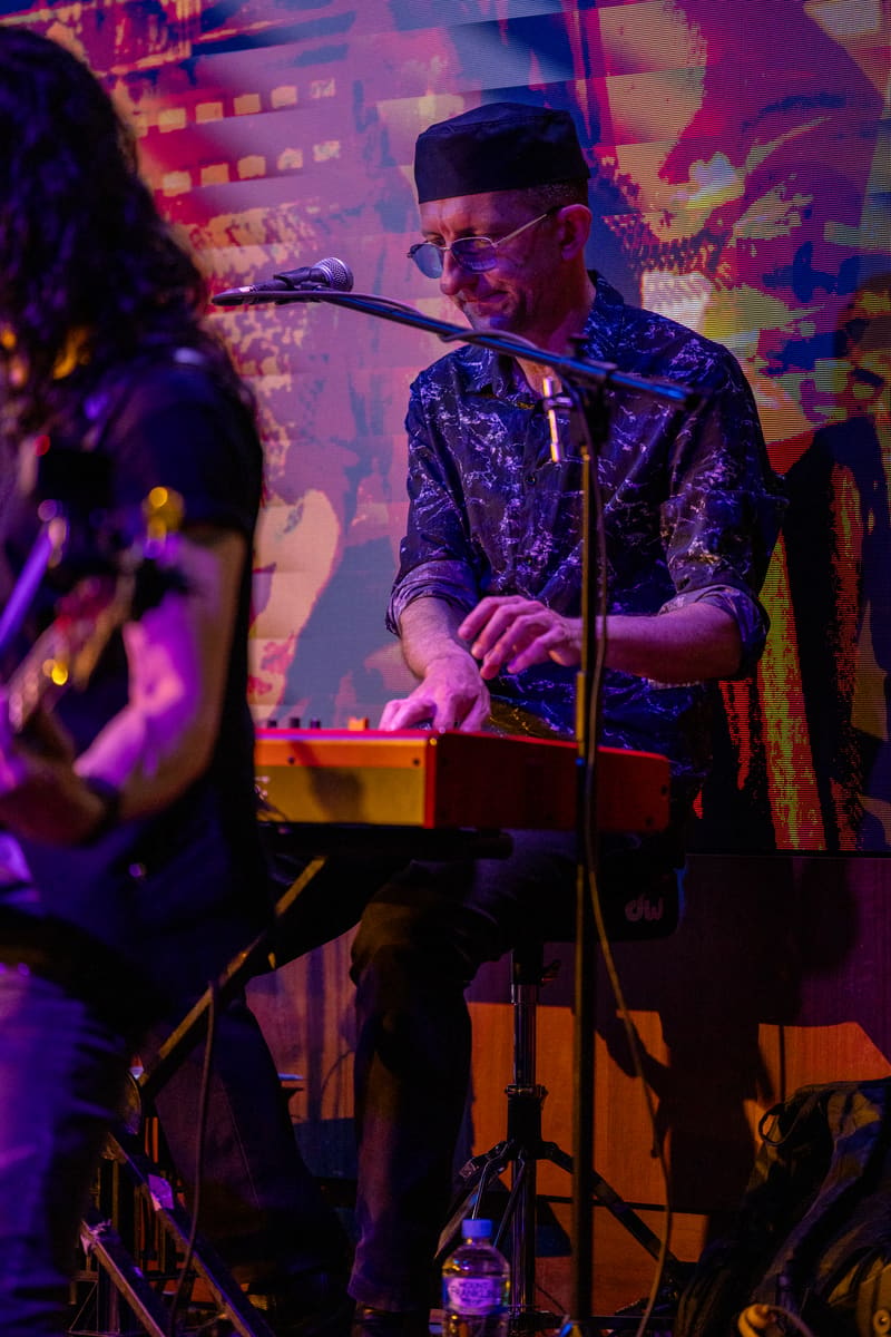 Musician in patterned shirt and cap performs at keyboard under purple and pink stage lighting, with band members visible nearby.