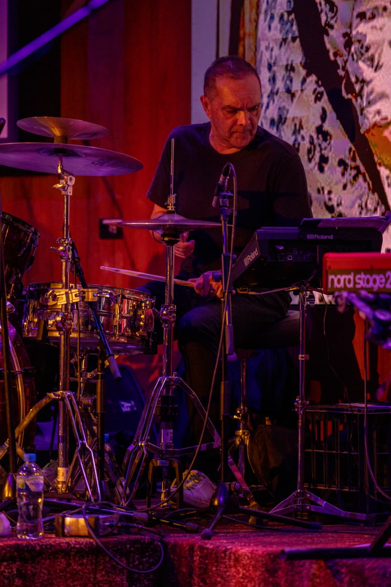 Drummer performs at an electronic drum kit on stage lit by purple and red spotlights.
