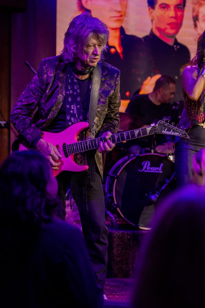 Guitarist in sequinned jacket performing with pink electric guitar under purple stage lighting, band members visible behind.