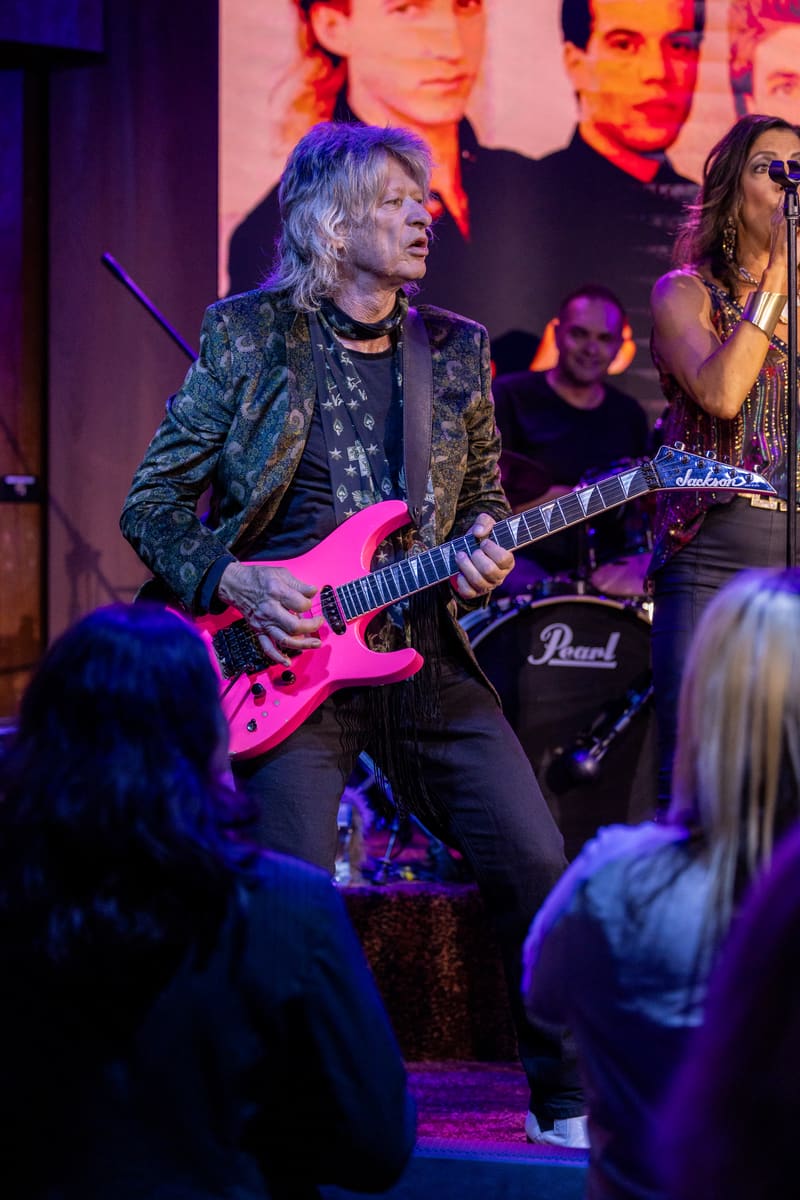 Guitarist in patterned jacket plays bright pink electric guitar under purple stage lighting with band members and audience visible.