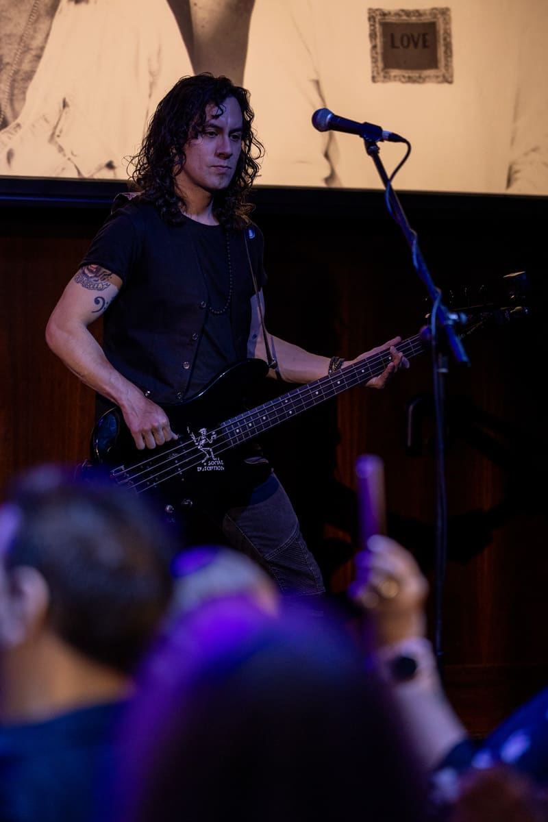 Musician with long dark hair playing bass guitar at microphone under purple stage lighting, audience in foreground.