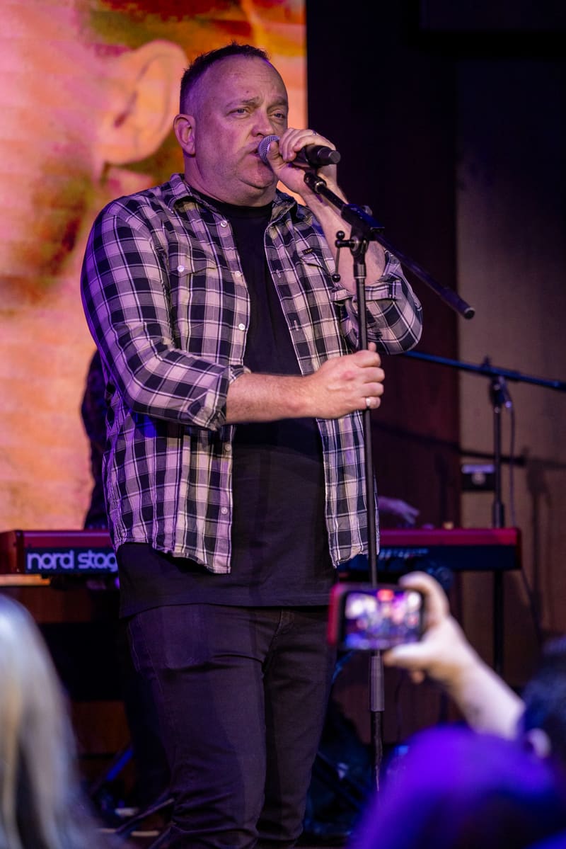 Male performer in purple-lit plaid shirt singing at microphone on stage during live music performance.