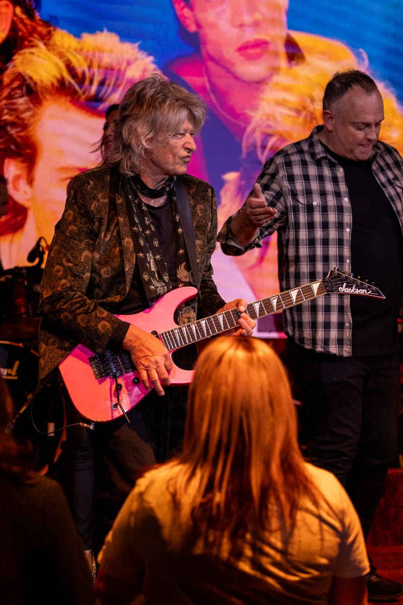 Guitarist in gold jacket performs with band member in checked shirt under colourful stage lighting, audience member visible in foreground.