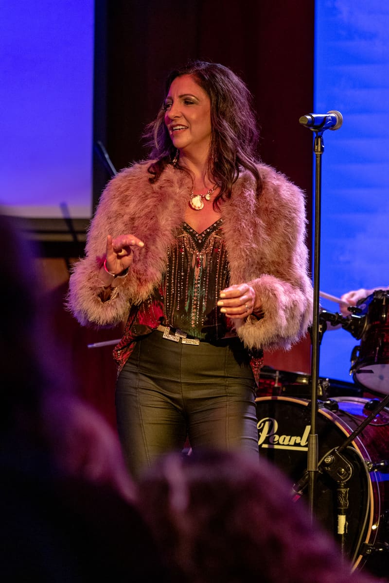 Performer in a fuzzy grey coat and dark outfit stands at a microphone on a stage lit with blue and purple lighting.