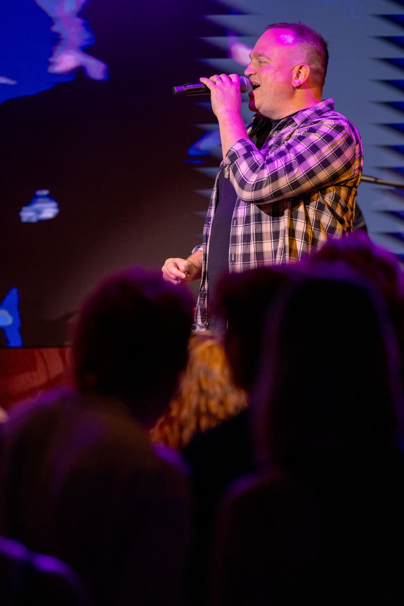 Singer in purple and white checked shirt performing at microphone beneath magenta stage lighting, audience silhouettes in foreground.