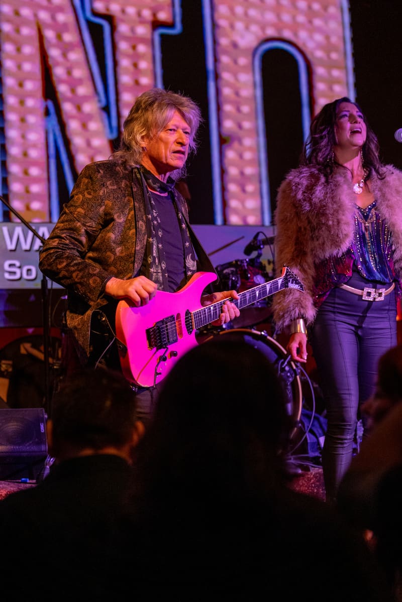 Musician plays hot pink electric guitar on stage beside a backing vocalist, illuminated by purple and pink stage lighting.
