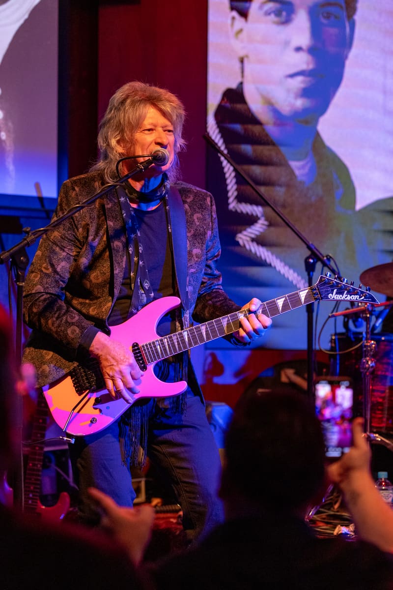 Musician performing with electric guitar under blue and pink stage lighting, with projected image on screen behind.