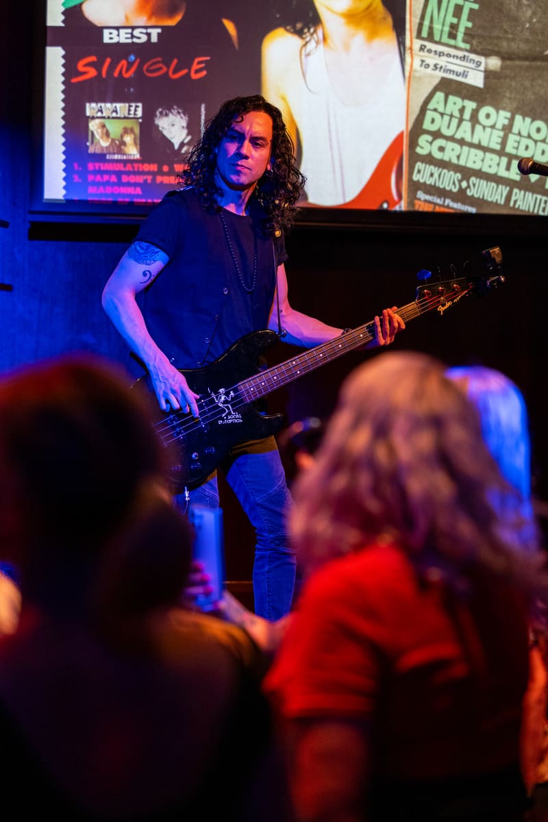 Bassist with long dark hair performs on stage beneath colourful signage as audience members watch from the foreground.