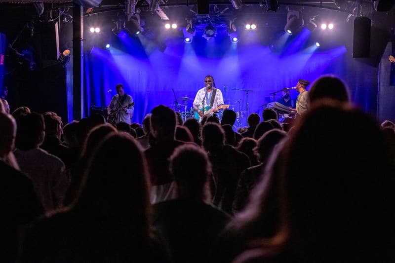 The Original Wailers perform on stage bathed in blue lighting, surrounded by an engaged crowd.