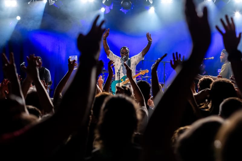 Performer on stage with arms raised amid a crowd with raised hands, lit by blue stage lighting.