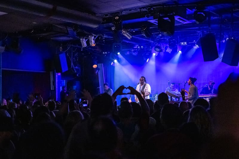 Band performing on stage bathed in blue and purple spotlights, with audience members raising phones and hands in the foreground.