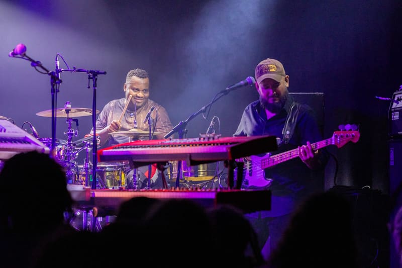 Two musicians perform on stage with keyboards and bass guitar under purple stage lighting during a live concert.