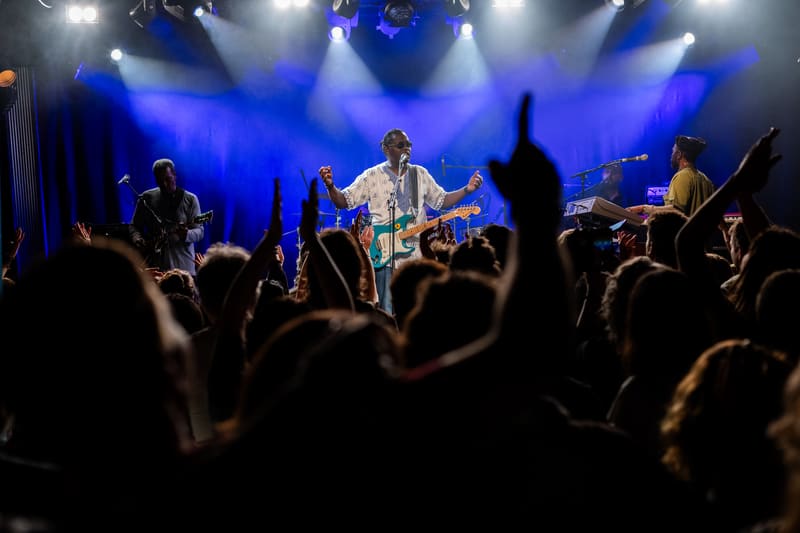 Musician in light-coloured shirt performing centre stage under blue spotlights whilst crowd raises hands.