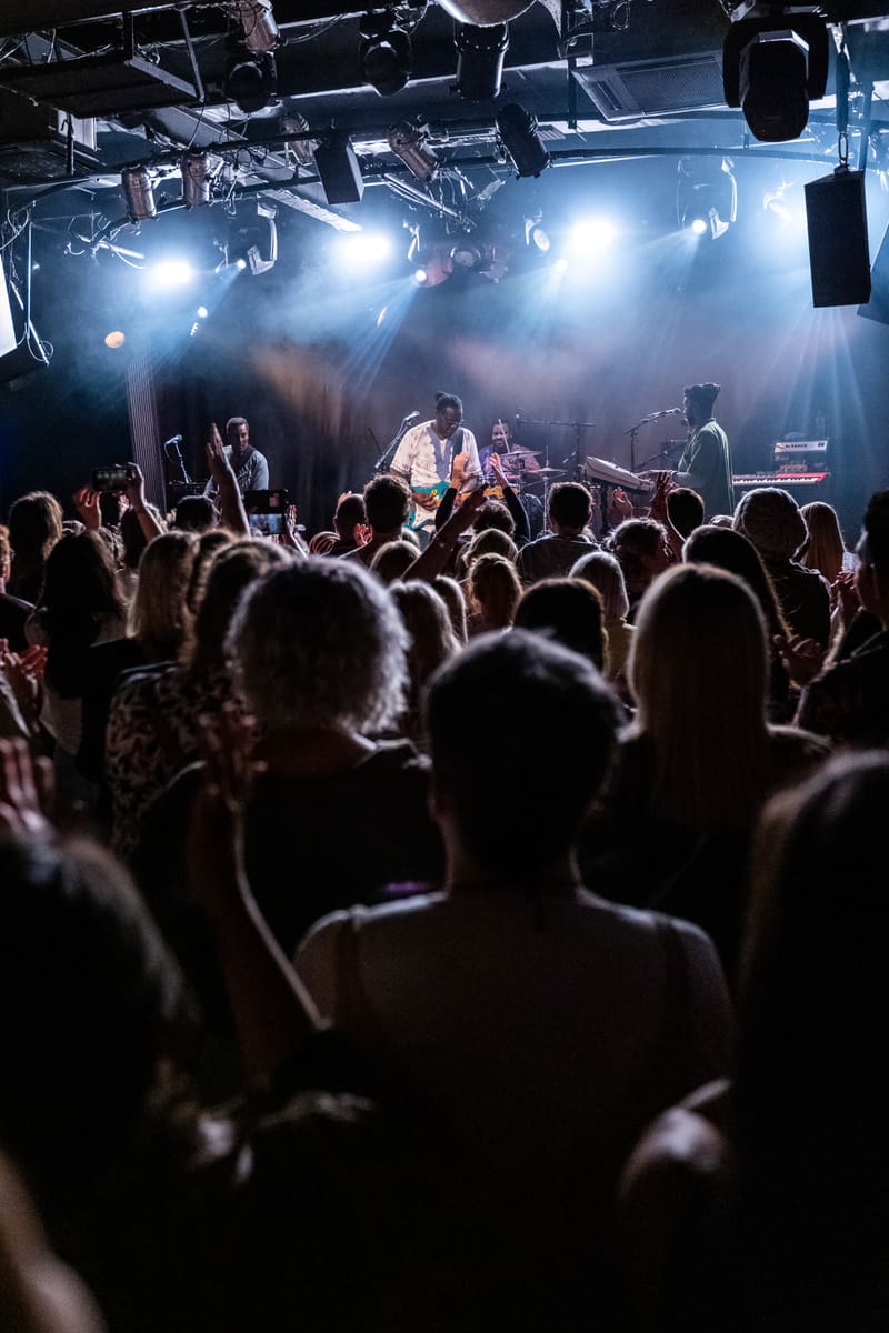 Crowd of concertgoers watching performers on a brightly lit stage under professional lighting.