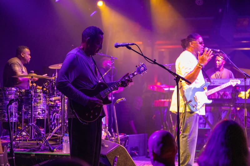 Band members perform on stage bathed in purple and gold lighting, with guitarist centre-stage at microphone and drummer visible left.