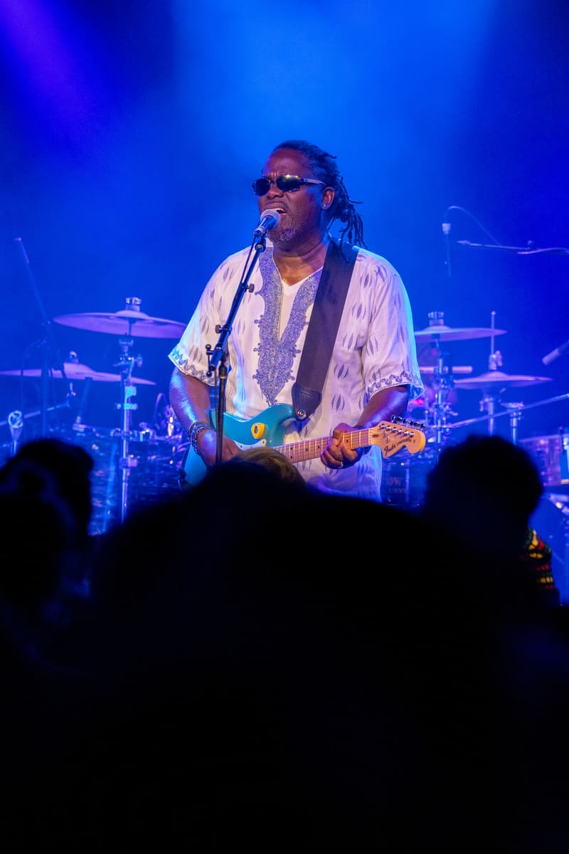 Guitarist performs on stage under blue lighting with audience silhouettes in foreground and drum kit visible behind.