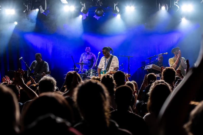 Five musicians perform on a blue-lit stage whilst a crowd of raised hands fills the foreground.