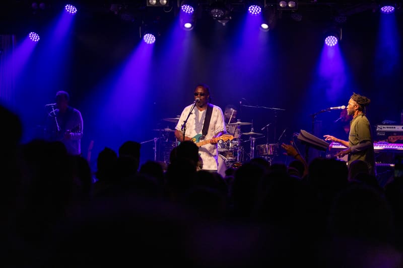 Musicians perform on stage bathed in blue spotlights whilst silhouetted audience members watch from below.