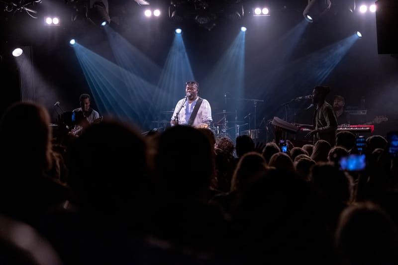 Vocalist in white shirt centre stage amid blue stage lights with silhouetted audience members in foreground.