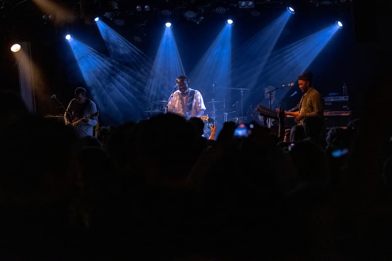 Five musicians perform on stage under blue spotlight beams during The Original Wailers concert.