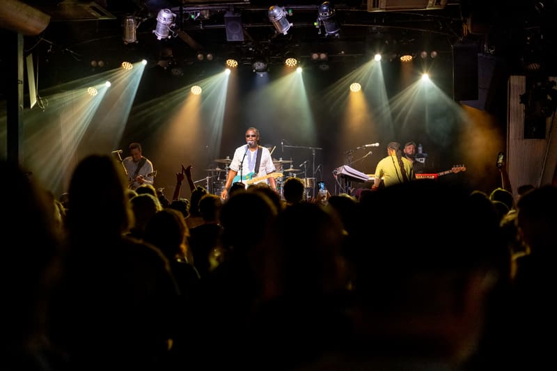 Band performs on stage under dramatic spotlights with audience silhouettes in foreground.